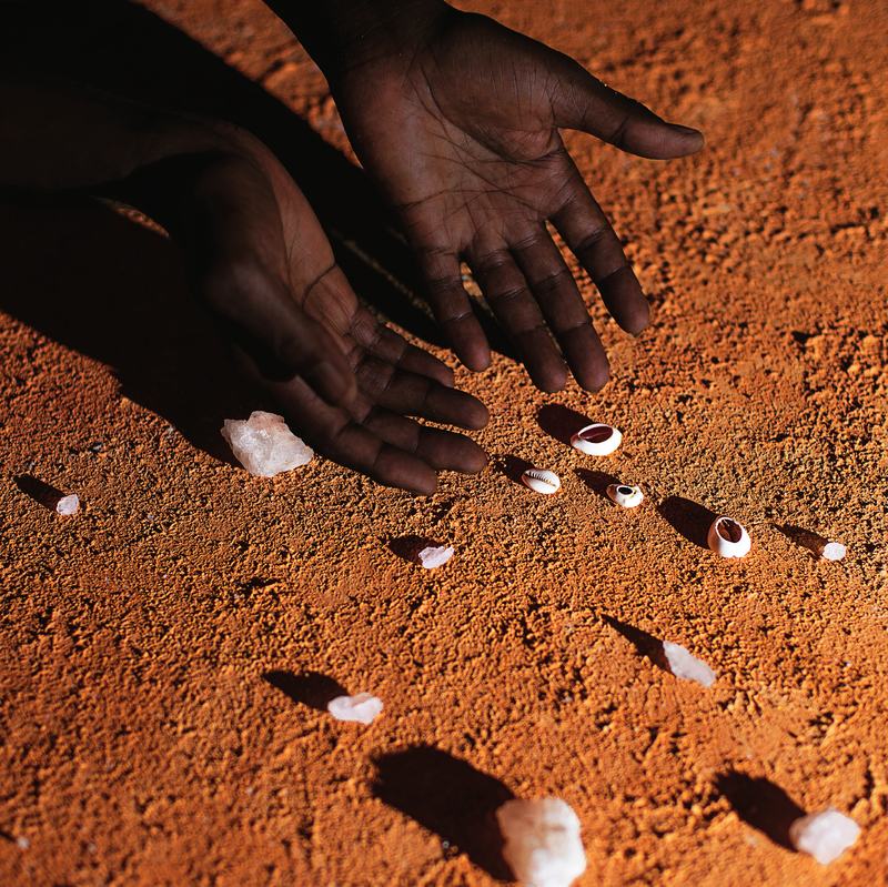 Open hands over cowrie shells and salt cast on red earth.